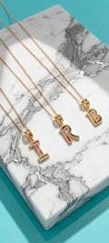 Three gold initial necklaces with letter pendants (T, R, B) decorated with colorful rhinestones, arranged on a white and gray marble tray over a turquoise background.