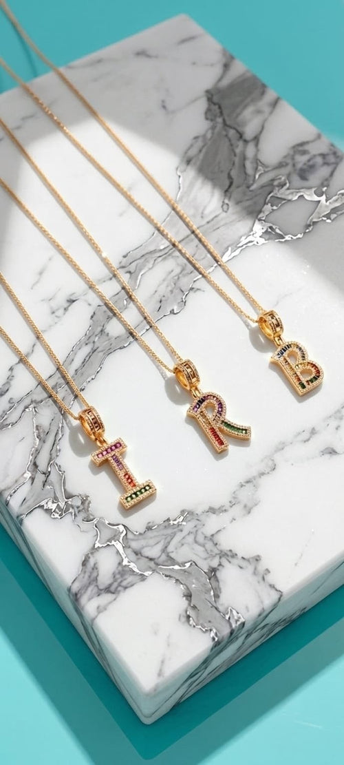 Three gold initial necklaces with letter pendants (T, R, B) decorated with colorful rhinestones, arranged on a white and gray marble tray over a turquoise background.