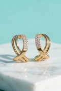 A pair of gold hoop-style earrings with pavé-set clear stones, displayed on a white marble surface against a light aqua background.