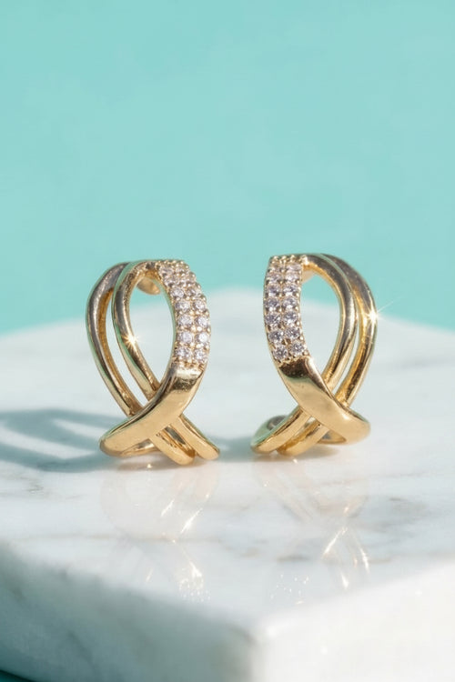 A pair of gold hoop-style earrings with pavé-set clear stones, displayed on a white marble surface against a light aqua background.