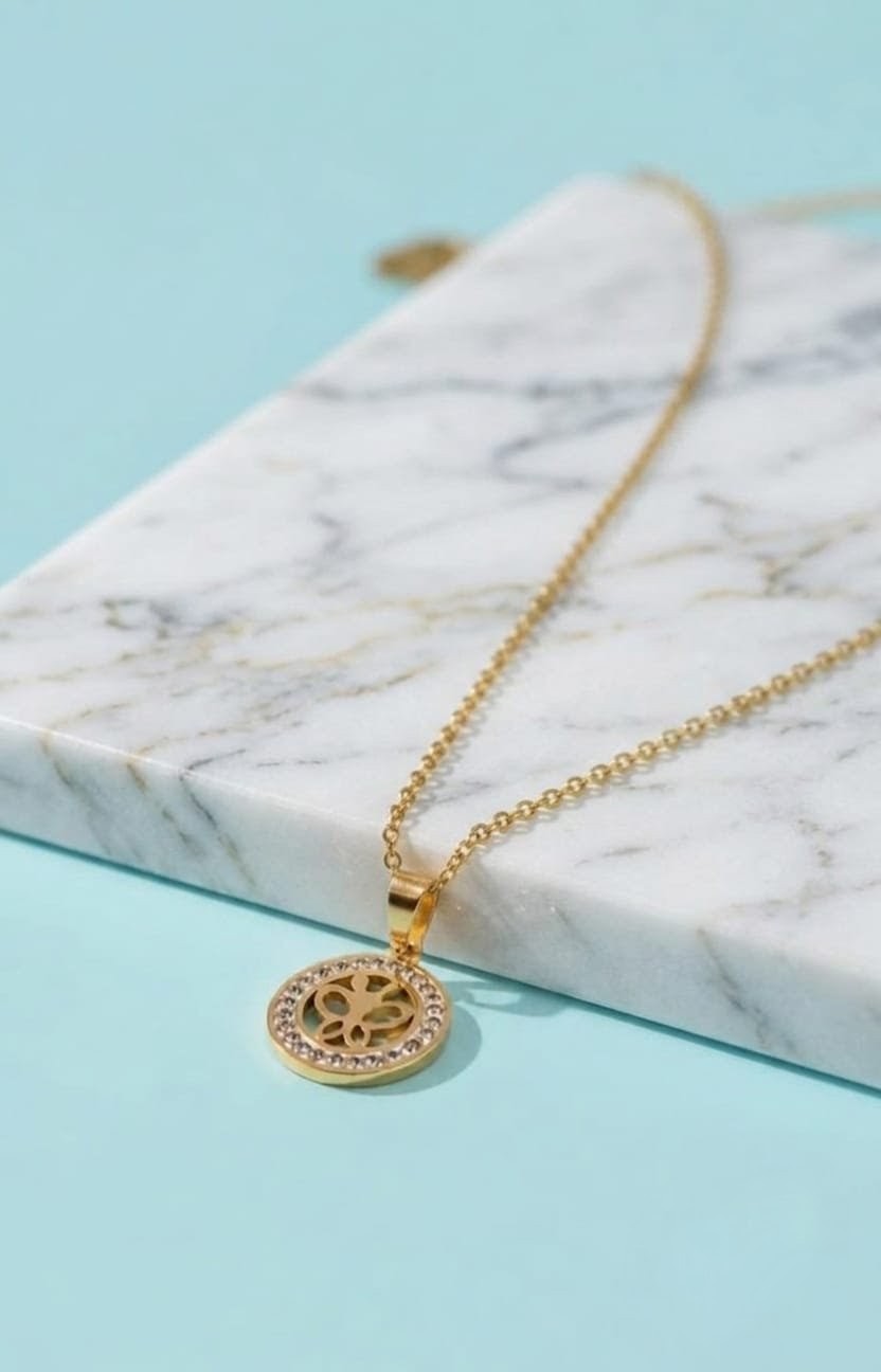 A gold pendant necklace with a circular sunburst design set with small clear stones, resting on a white marble block against a light blue background.